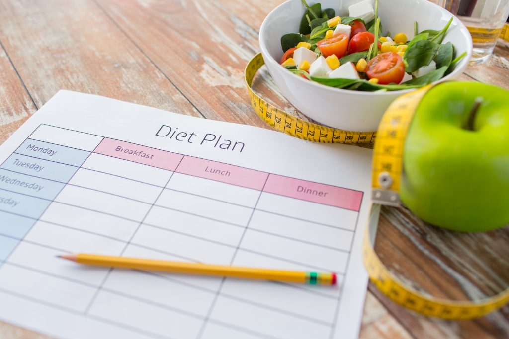 A nutritionist or dietitian’s paper diet plan on a desk along with a pencil, apple, tape measure and salad in a white bowl