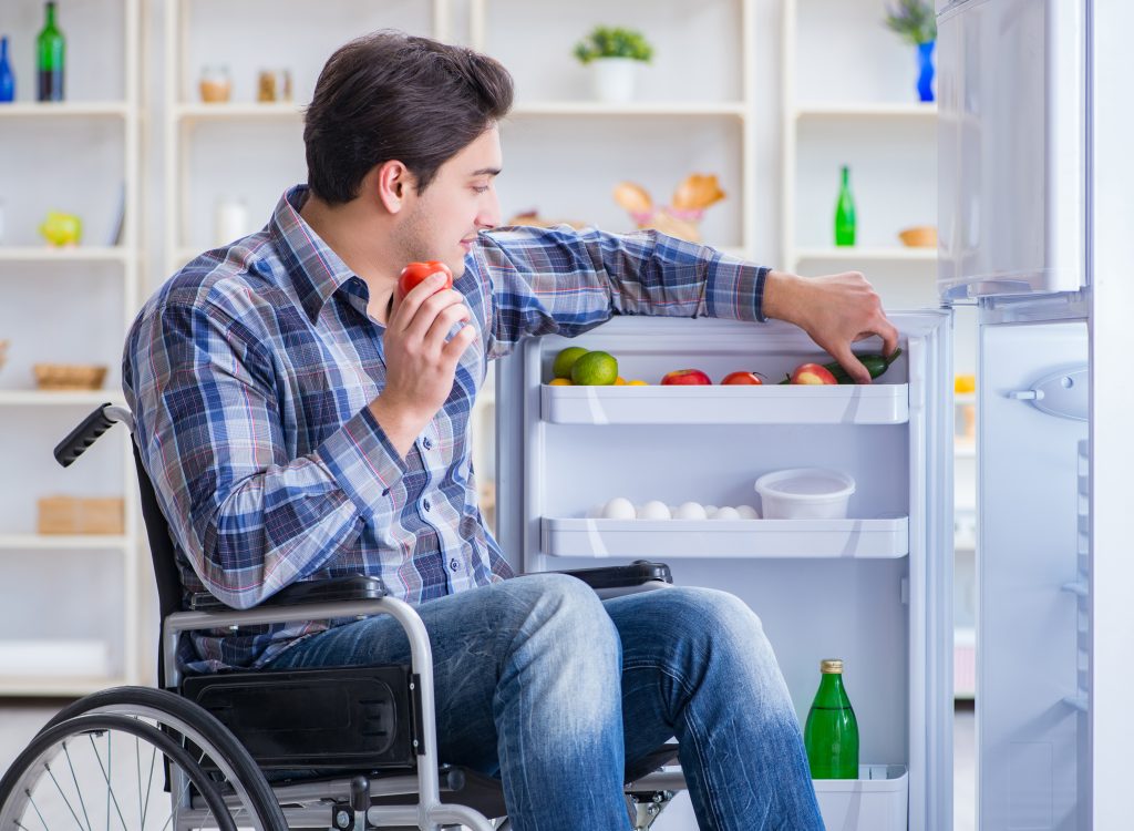 Young man with disability seated in a wheelchair beside an open fridge door, choosing healthy foods to eat
