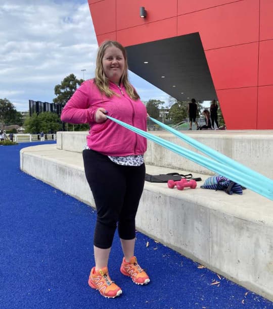 Image of Bonnie using a standing row exercise with added resistance via a blue theraband to build her strength and posture to reach her NDIS goals