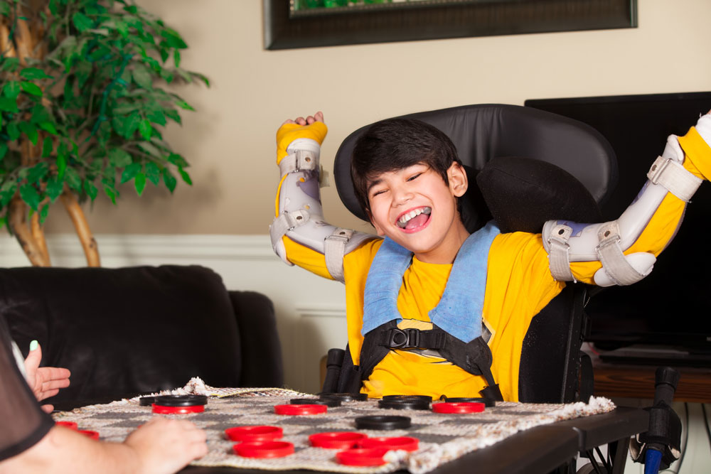 Boy with cerebral palsy seated in wheelchair with splints on his arms, playing a game of checkers.