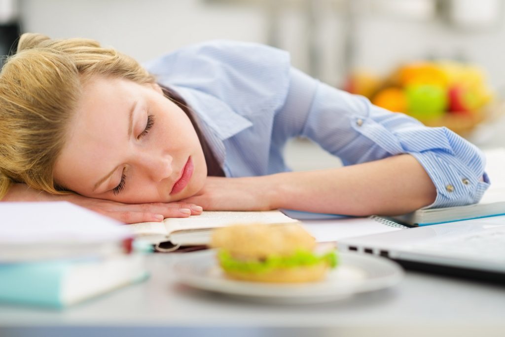 A young woman with chronic fatigue asleep at her desk with an uneaten sandwich in front of her.