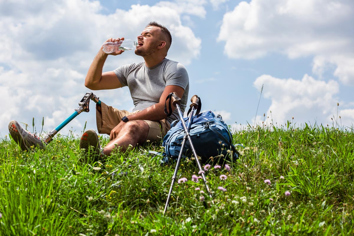 Young man with a prosthetic leg sitting in the grass and drinking water.