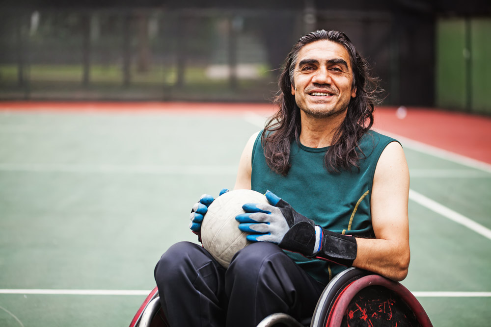 Man with a spinal cord injury seated in a wheelchair on a court holding a ball, ready to play wheelchair sport.