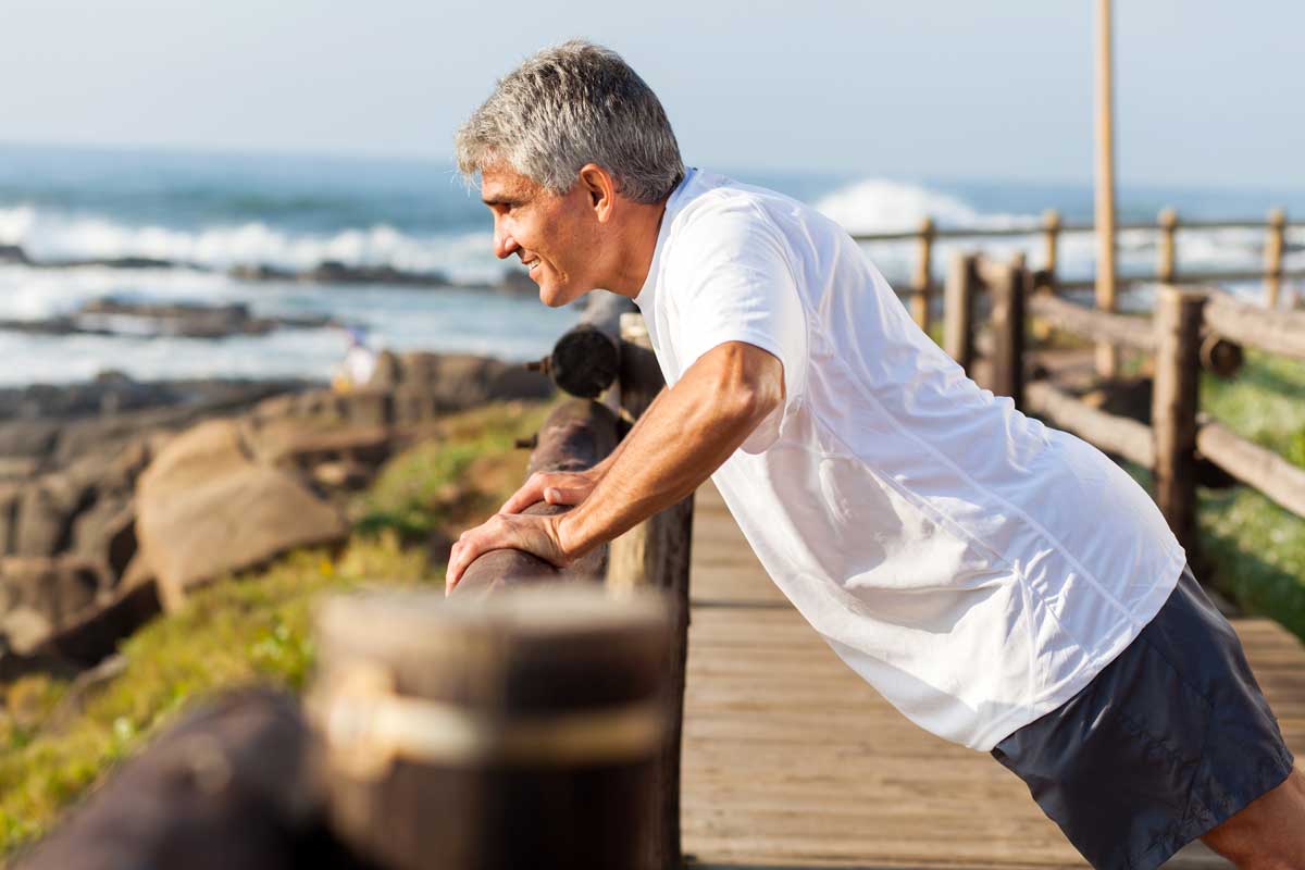 Older man with grey hair doing push up exercise beside the beach. 