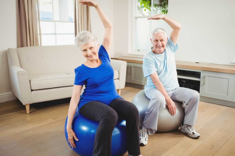Older man and woman both sitting on exercise balls in their home and performing stretching exercises.
