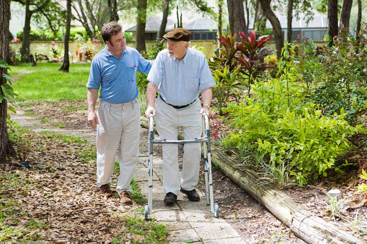 Physiotherapist walking through a garden with an older man using a walking frame.