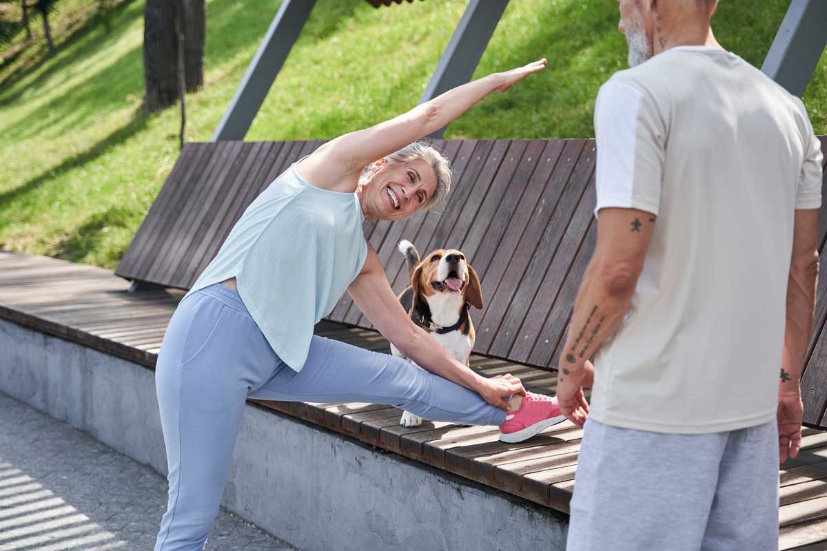 Older woman wearing a T-shirt and exercise pants stretching her leg on a park bench, accompanied by a man and a beagle.