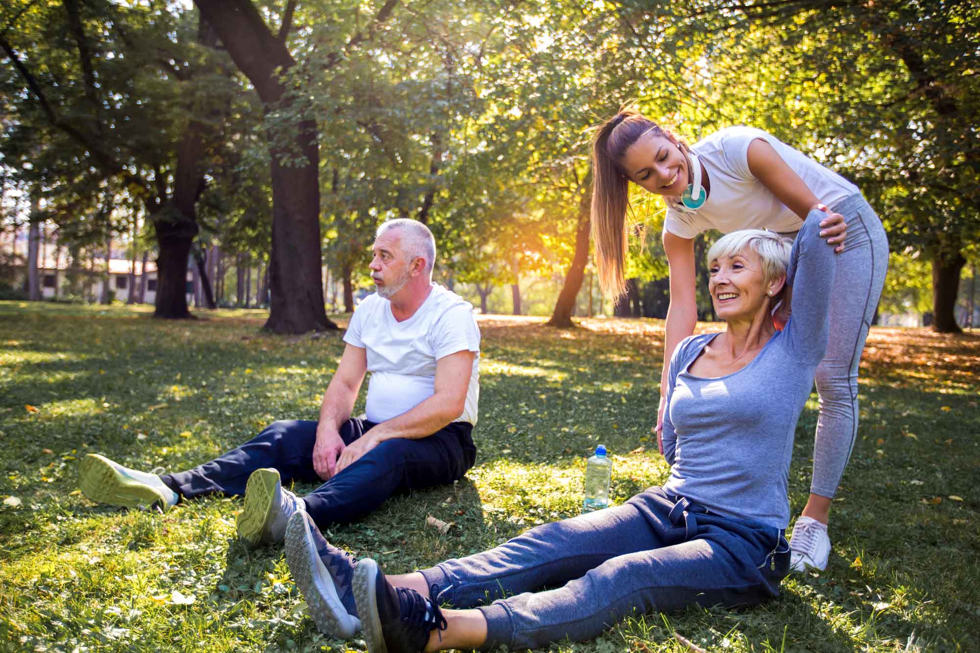 An older man and woman sitting on the grass in a park, with a physiotherapist helping the woman stretch her arms.