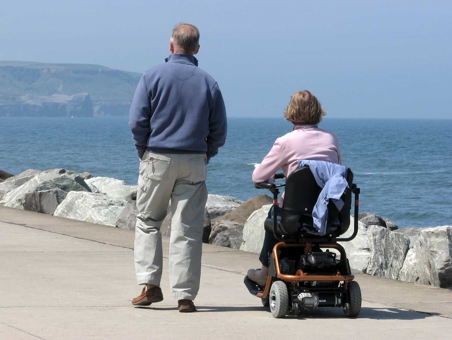 Woman with motor neuron disease using a motorized wheelchair along a waterside pathway, accompanied by a man walking beside her. 