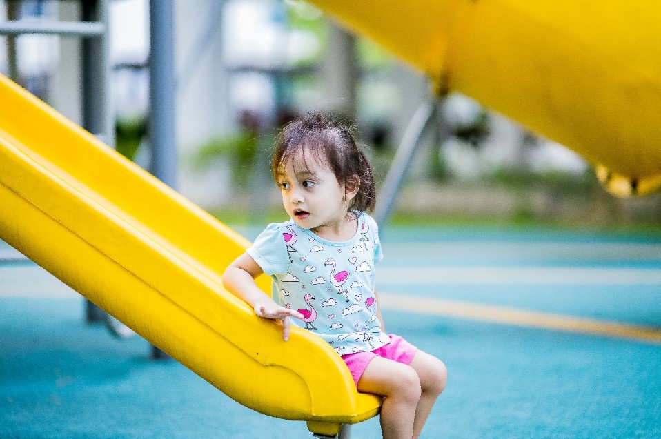 young girl with brown hair who has developmental delay sitting on the end of a bright yellow slide.
