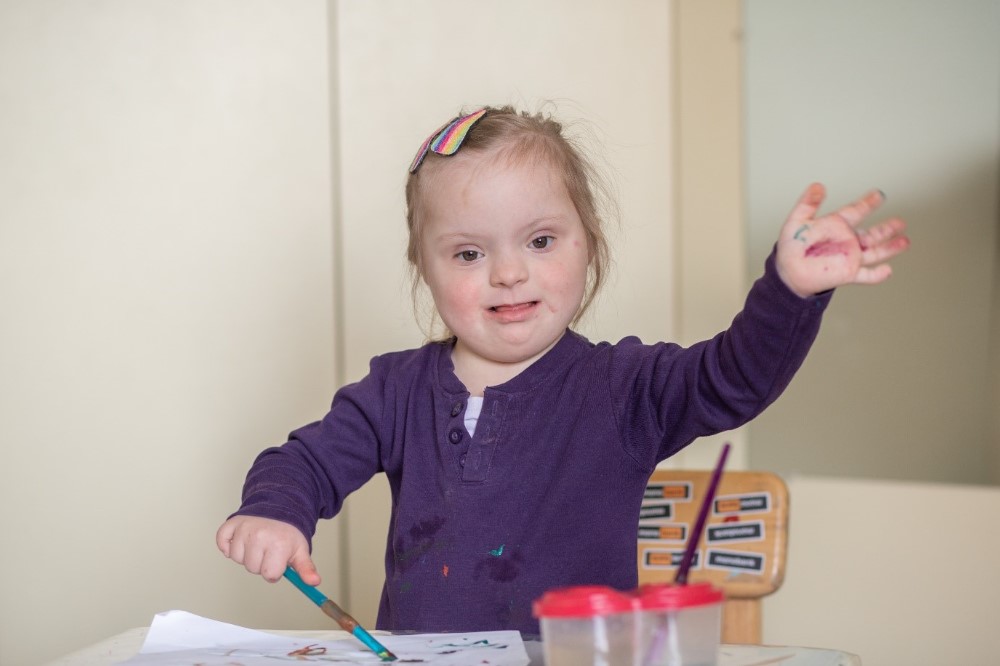 Young blond girl with global developmental delay related to Down syndrome seated at a table painting a picture.