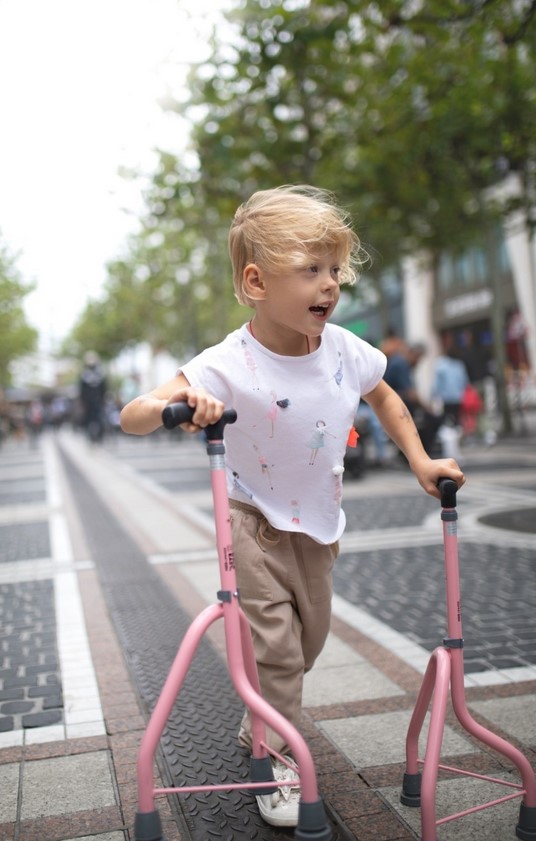 A young child with a paediatric mobility condition walking with pink modified crutches along a paved street. 