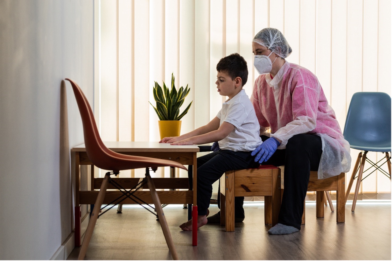 Woman wearing personal protective equipment conducting a paediatric physiotherapy assessment on a boy with disability sitting on a stool at a table.  