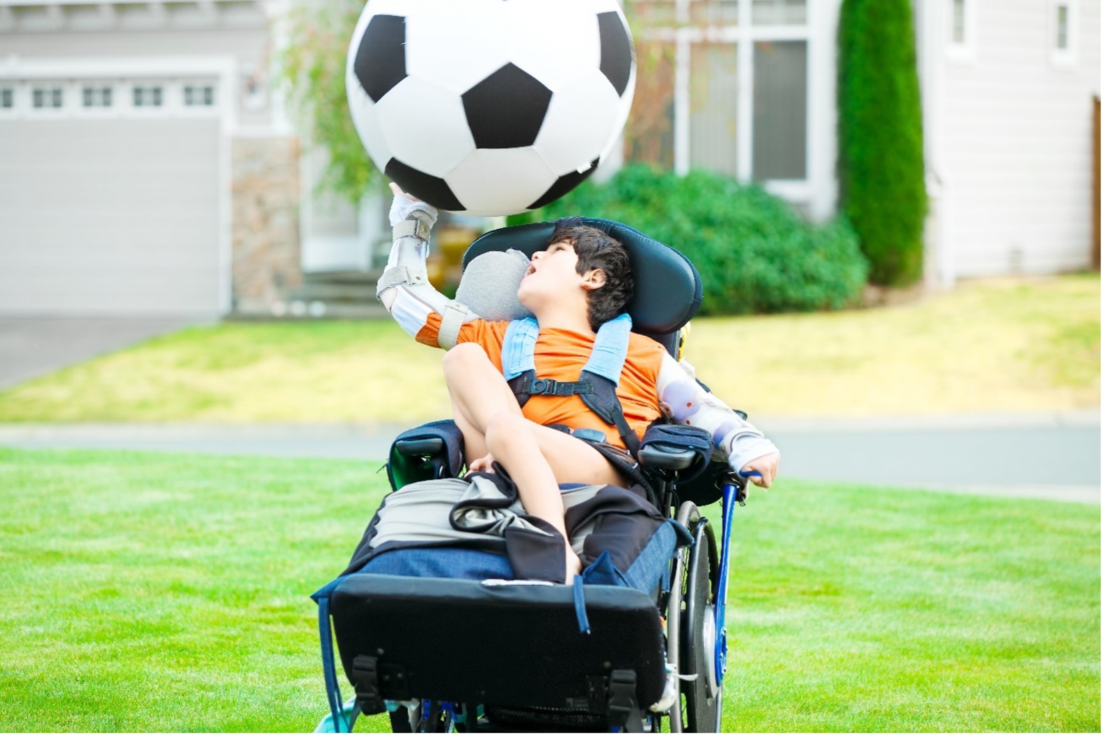 A young boy with disability seated in a blue wheelchair playing with an oversized soccer ball. 