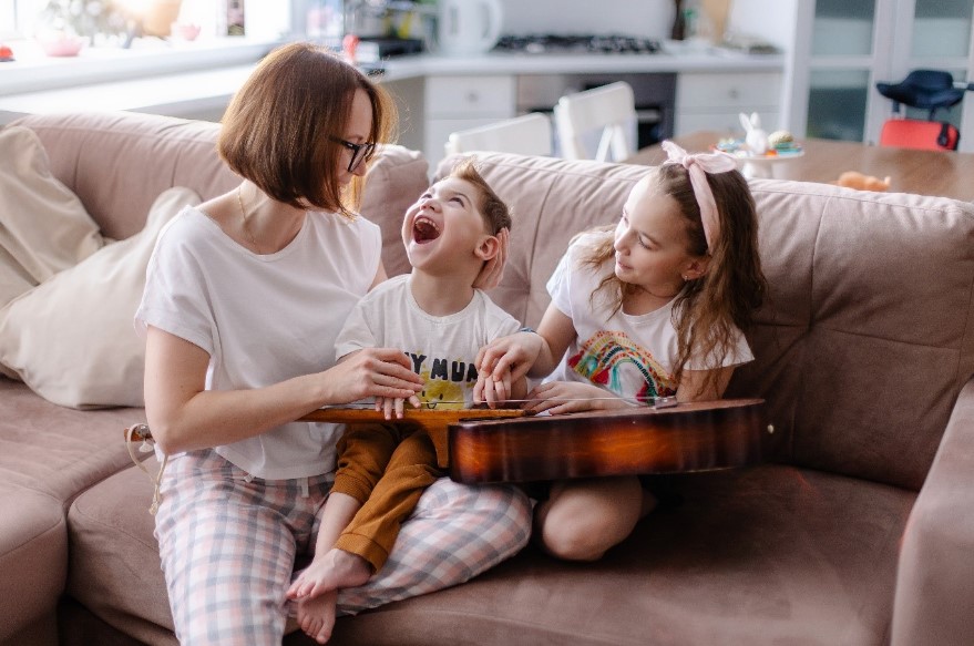 A young child with disability sitting on a couch with his mum and sister doing finger exercises on a guitar.