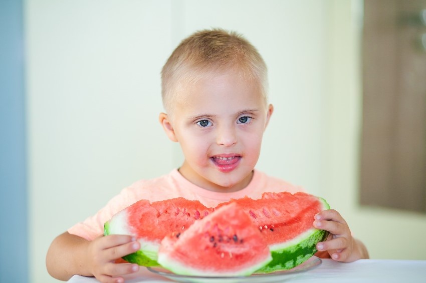 A young child with disability standing at a table in front of a plate of cut watermelon. 