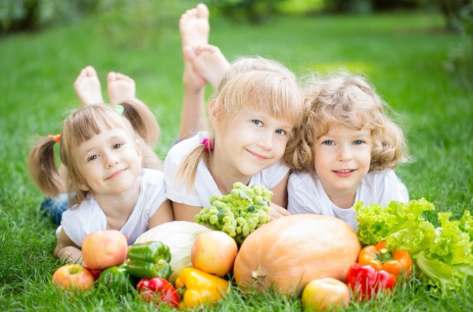 Three smiling young children lying on the grass with a variety of fresh colourful fruits and vegetables in front of them
