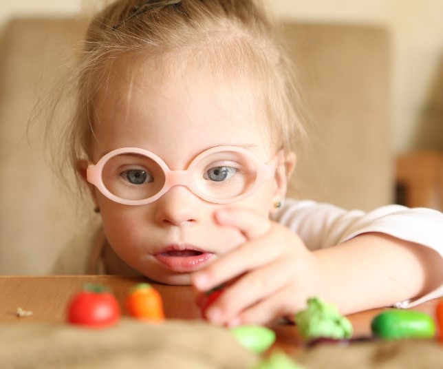 A young girl with disability wearing pink glasses is looking at and touching different vegetables on a table in front of her