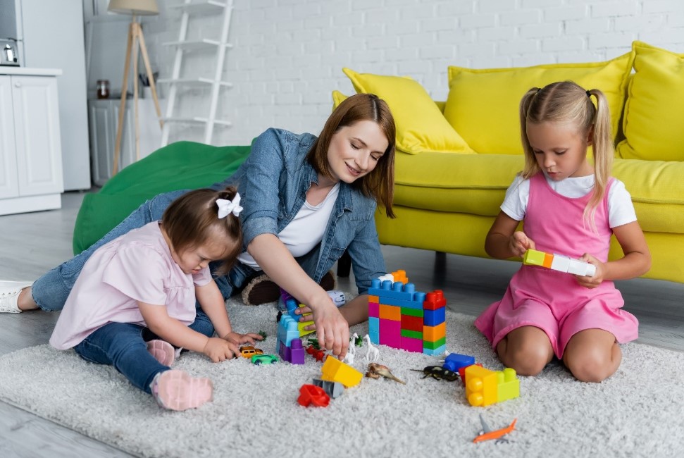 Two young girls with developmental delay sitting on a rug doing play-based physiotherapy using colourful blocks and toy cars and animals.