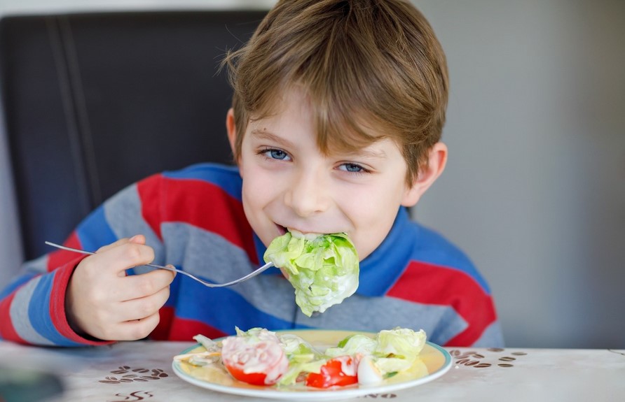 A young boy in a striped shirt sits at a table eating a plate of salad.