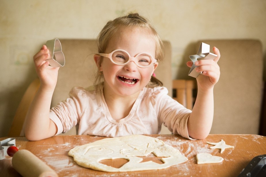 A young girl with a disability wearing a pale pink shirt and glasses sits at a table cutting cookie shapes out of dough.