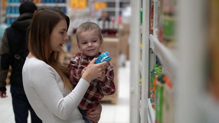 A young mother stands in the supermarket aisle holding a young boy. They are looking at the label on a juice popper to check for chemicals that can cause a food allergy in children.