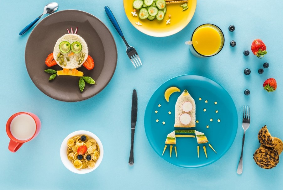 Birds eye view of several serves of kids’ food placed on a table. Food has been served on colourful plates and arranged in fun shapes, including a rocket and a fish shape.