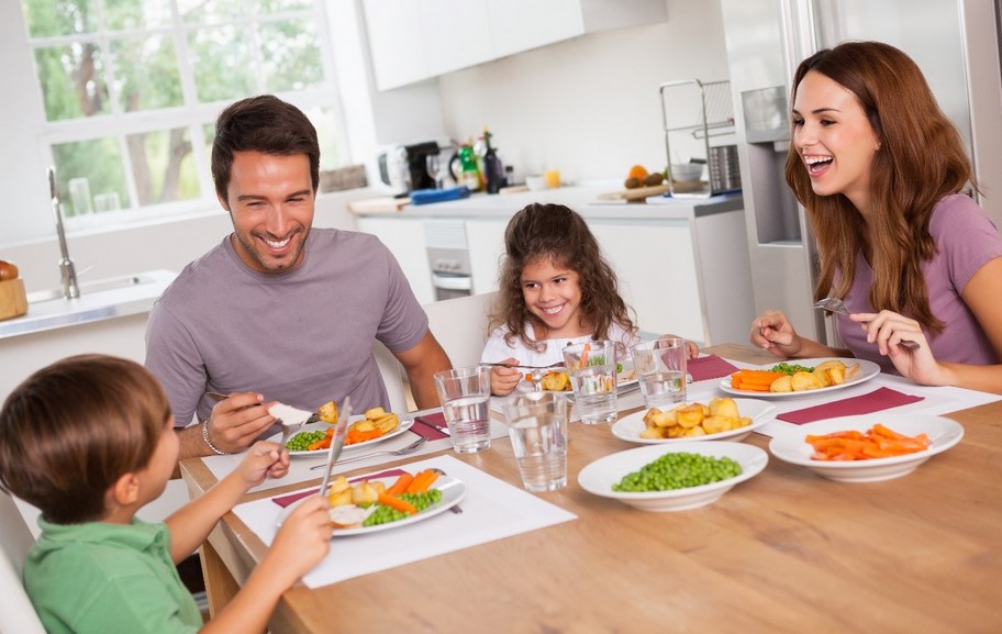 A family of four (mum, dad, boy and girl) sit at a table eating a healthy meal of chicken and vegetables. They are all smiling and look like they are enjoying a relaxed healthy meal together.