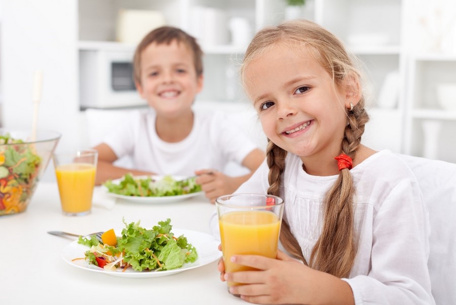 A smiling young girl and boy both dressed in white tops are sitting at a table with plates of salad and glasses of orange juice in front of them.