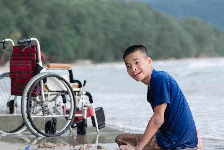 A boy with disability wearing a blue shirt sits on the sand at the water’s edge on a beautiful beach with his wheelchair and toys beside him.