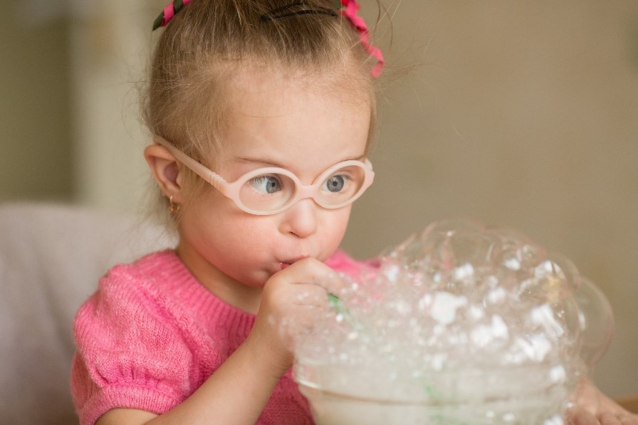 A young girl with disability wearing a pink top is doing an early intervention therapy activity blowing bubbles through a straw.