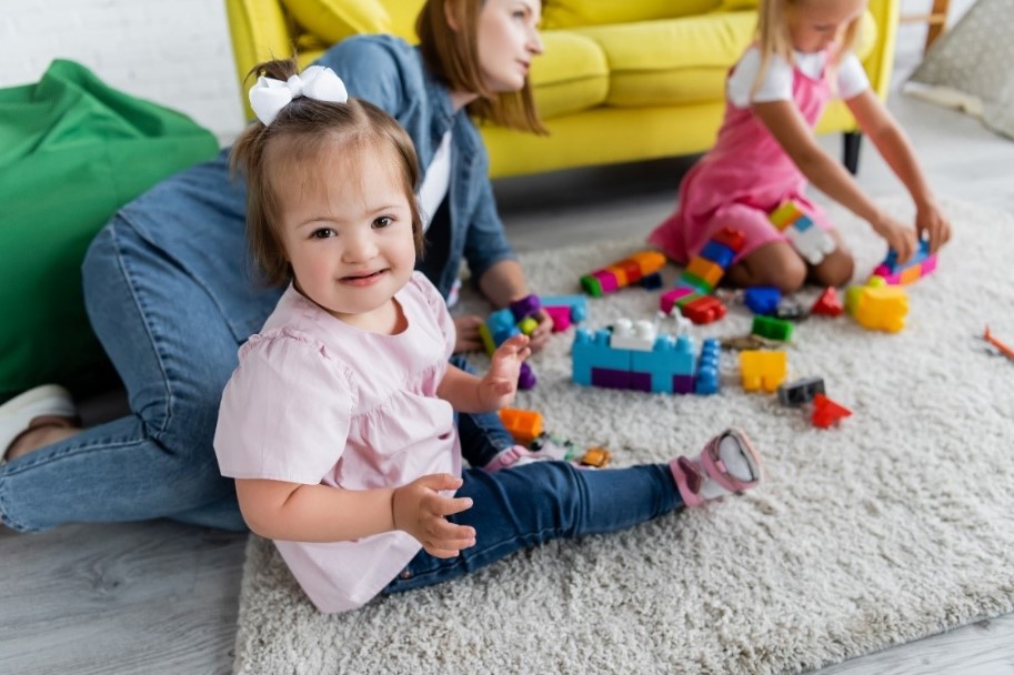 A young girl with disability sits on a rug on the floor doing play-based early childhood intervention activities with blocks.