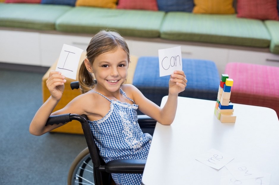 A young girl with disability sits in a wheelchair at a desk holding cards that say ‘I can’.