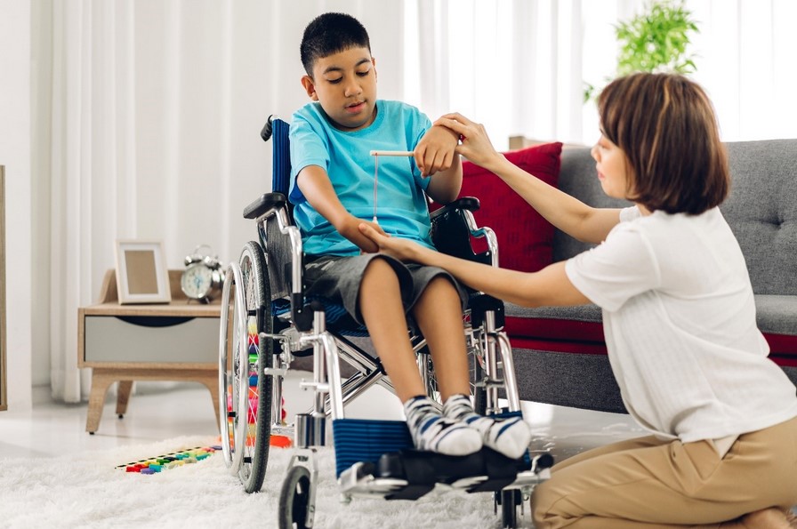 A boy with disability wearing a blue shirt and sitting in a wheelchair is doing arm and hand exercises with a physiotherapist who is kneeling on the floor beside him.