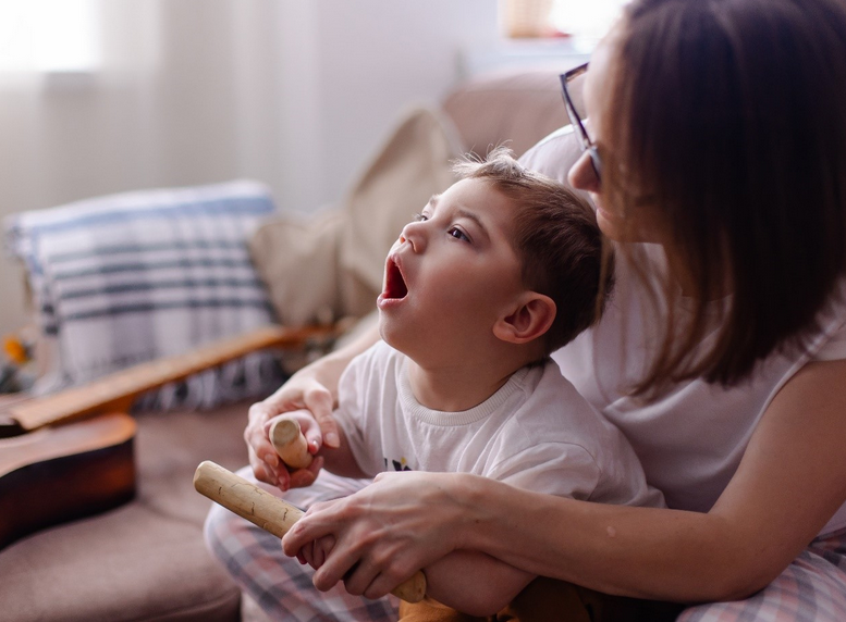  A very young boy with cerebral palsy sits on his mother’s lap. She is helping him to make music with rhythm sticks. 