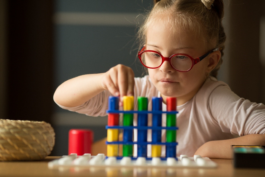 A young blond-haired girl with disability wearing red glasses sits at a table. She is stacking colourful blocks on top of each other on a board.